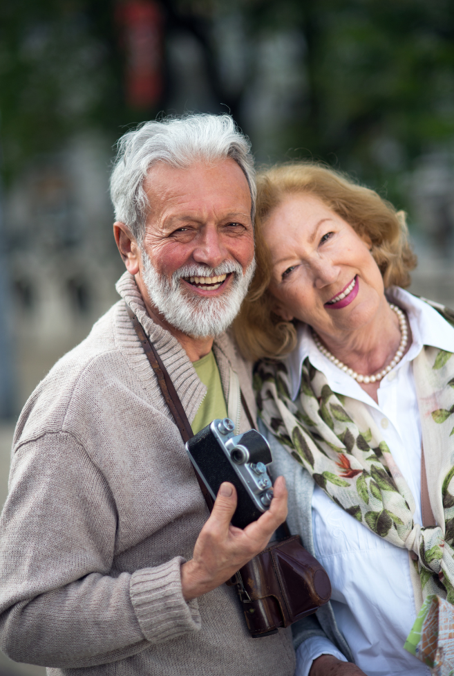 A smiling elderly couple, one holding a camera, standing close together in a display of companionship.