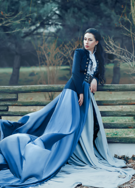 An individual in an elegant blue and black dress stands beside a wooden bench, with a serene park setting in the background.