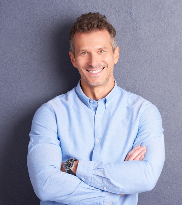Health Coverage Confidence A smiling, confident individual with crossed arms wearing a blue shirt and a watch, standing against a gray background