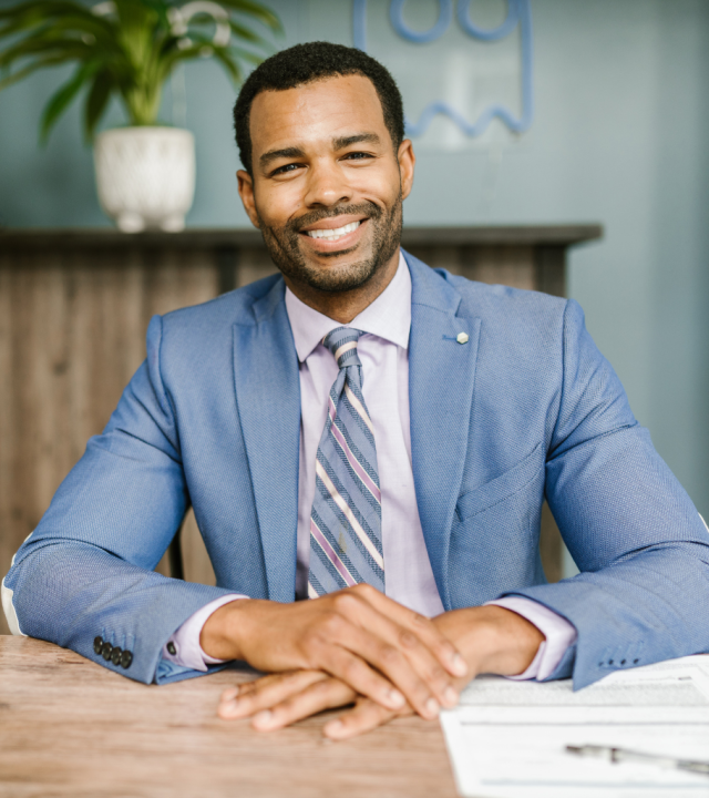 Professional Health Insurance Services for Diplomats A smiling professional looking into the camera, sitting at a desk with hands clasped, wearing a blue suit and striped tie, in an office setting with documents on the table