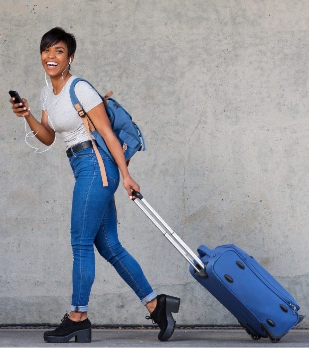 Stay Connected and Covered on the Go A person walking with a rolling suitcase and wearing a backpack, looking at a smartphone with earphones plugged in, symbolizing the convenience of having travel health insurance.